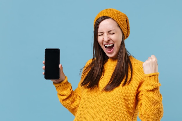 Menina morena jovem feliz no chapéu de suéter amarelo posando isolado sobre fundo azul. conceito de estilo de vida de pessoas. mock up espaço de cópia. segure o celular com tela vazia em branco fazendo gesto de vencedor.