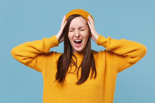 Menina morena jovem frustrada no suéter chapéu posando isolado no retrato de estúdio de fundo azul. Conceito de estilo de vida de pessoas. Mock up espaço de cópia. Gritando, mantendo os olhos fechados, coloque as mãos na cabeça.