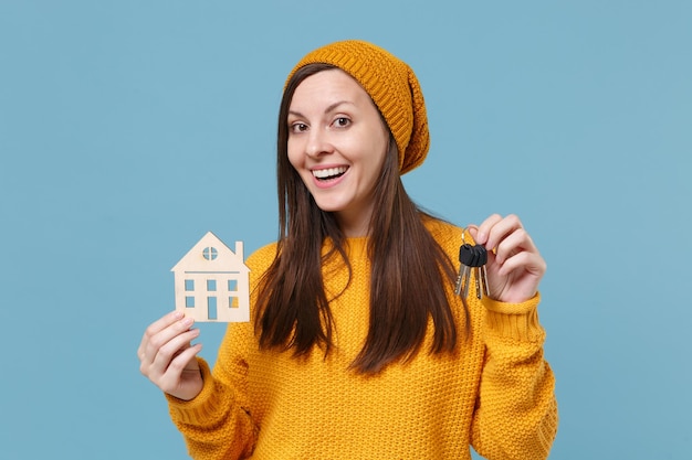 Menina morena jovem sorridente na camisola amarela e chapéu posando isolado no retrato de estúdio de fundo azul. Conceito de estilo de vida de emoções sinceras de pessoas. Mock up espaço de cópia. Segure o molho de chaves da casa.