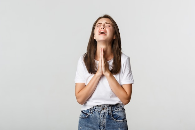 Foto menina morena miserável de mãos dadas, orando e implorando, implorando por ajuda