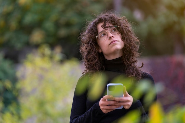 Menina morena pensando no que digitar em seu telefone inteligente no parque com algumas árvores ao fundo