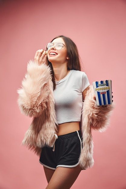 Menina morena rindo com pelo rosa falso segurando pipoca na mão, vestindo shorts preto e blusa branca.