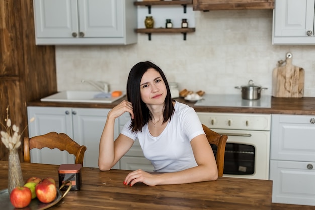 Menina morena se senta à mesa na cozinha olha para o quadro.