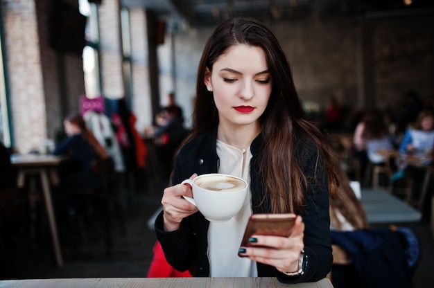 Menina morena sentada no café com uma xícara de cappuccino e olhando para o telefone móvel.