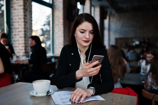 Menina morena sentada no café com uma xícara de cappuccino, ouvindo música em fones de ouvido e olhando para o telefone móvel.