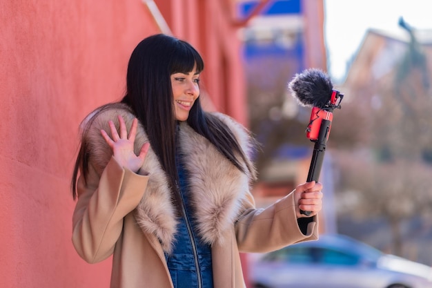 Menina morena sorrindo e acenando enquanto grava um blog de vídeo com seu celular e microfone, vestindo uma jaqueta de inverno para redes sociais. Caucasiano muito um fundo vermelho