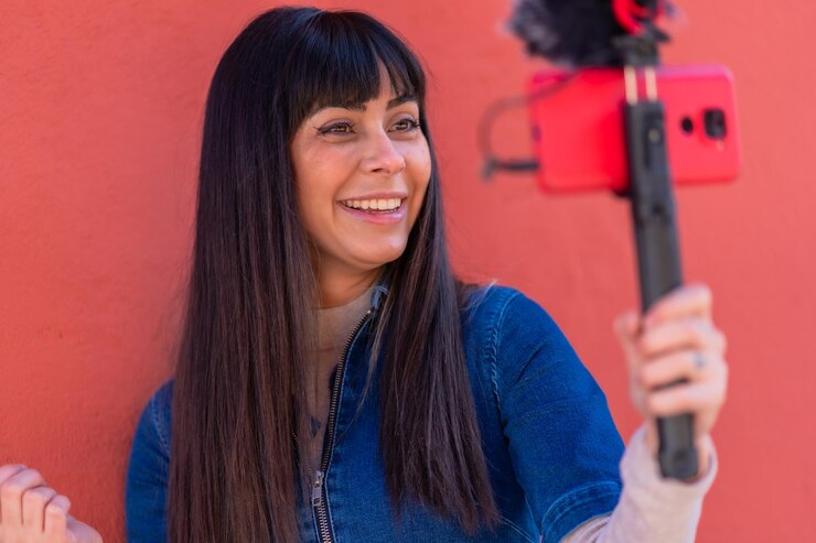 Foto menina morena sorrindo gravando um blog de vídeo com o celular e o microfone, vestindo um terno jeans azul para redes sociais. caucasiano muito um fundo vermelho
