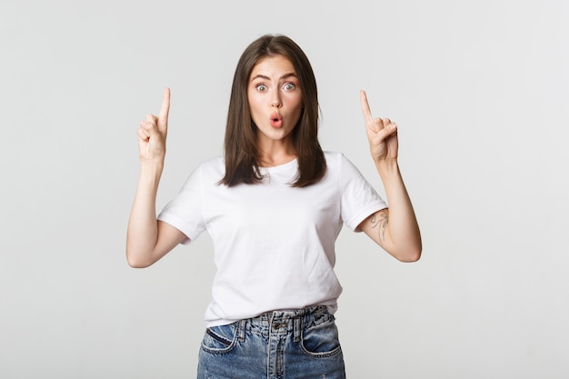 Menina morena surpresa e impressionada olhando espantada e apontando os dedos para cima.