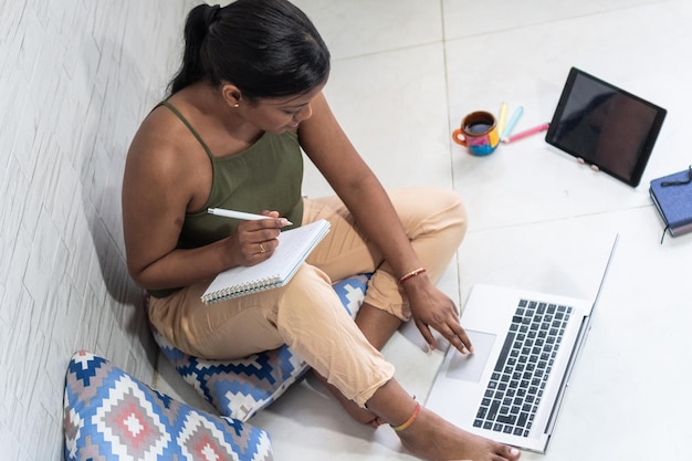 Menina morena tomando notas sentada no chão com laptop e café perto da janela