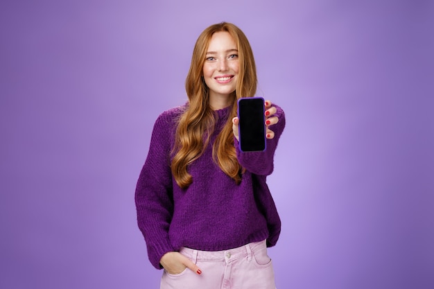 Menina mostra a tela do smartphone na câmera para pedir a opinião de um amigo sorrindo amplamente com uma expressão otimista e alegre, segurando a mão no bolso, promovendo o celular ou aplicativo sobre fundo roxo.