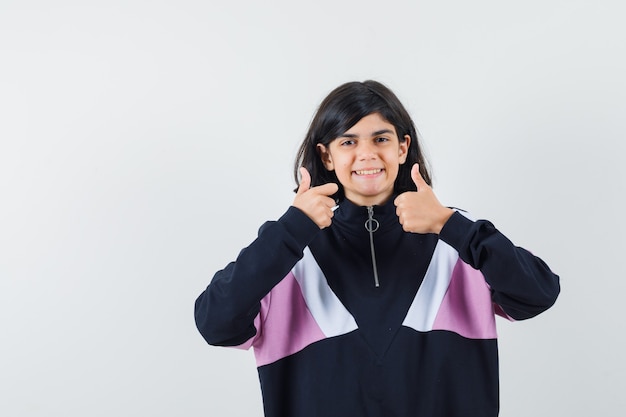 Menina mostrando dois polegares para cima na camisa e olhando feliz, vista frontal.