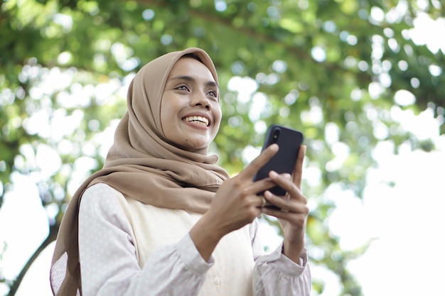 Menina muçulmana asiática em roupas casuais está usando um smartphone e sorrindo em pé na área externa