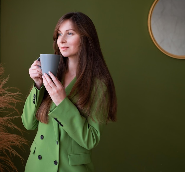 Menina muito adulta com cabelos castanhos compridos vestindo paletó verde bebendo chá ou café Jovem de negócios segurando caneca cinza