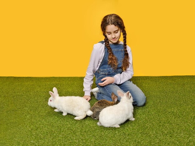 Menina muito bonitinha está deitada brincando com os coelhinhos no fundo amarelo do estúdio