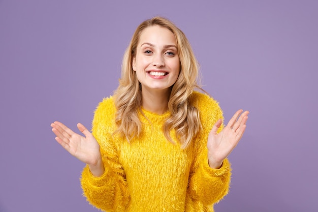 Menina muito jovem sorridente na camisola de pele amarela posando isolado no fundo da parede violeta pastel. Conceito de estilo de vida de emoções sinceras de pessoas. Mock up espaço de cópia. Espalhando as mãos apontando para o lado.