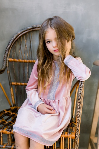 Menina muito pequena no lindo vestido rosa senta-se na cadeira de madeira e parece sonhadora, beleza e moda conceito, retrato interior