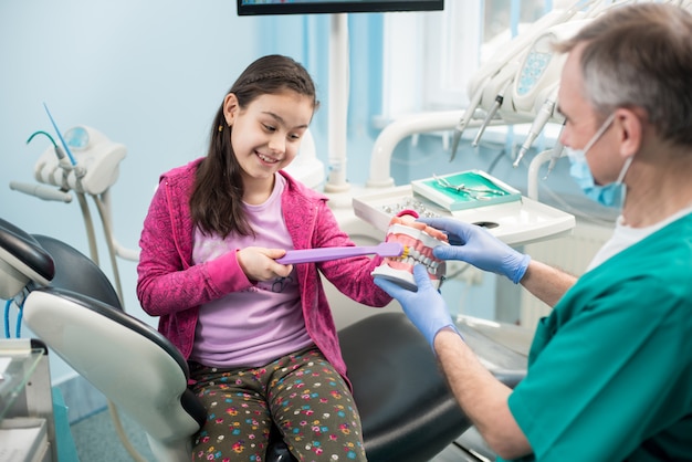 Menina na cadeira do dentista, educando a escovação adequada pelo dentista pediátrico, usando o modelo da mandíbula e a escova de dentes no consultório odontológico