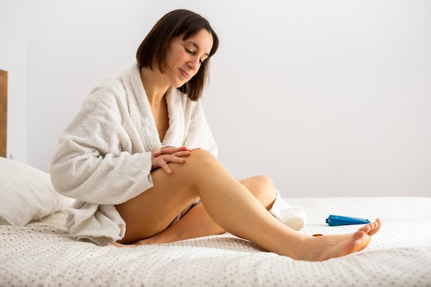 Menina na cama de roupão colocando creme nas pernas depois do banho