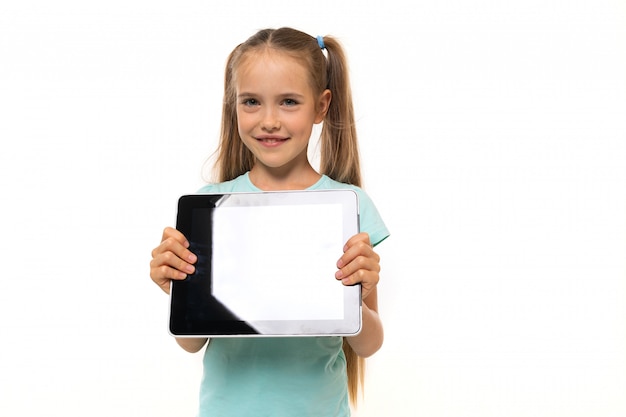 Menina na moda adolescente em camiseta azul com tablet gesticula e sorri na parede branca