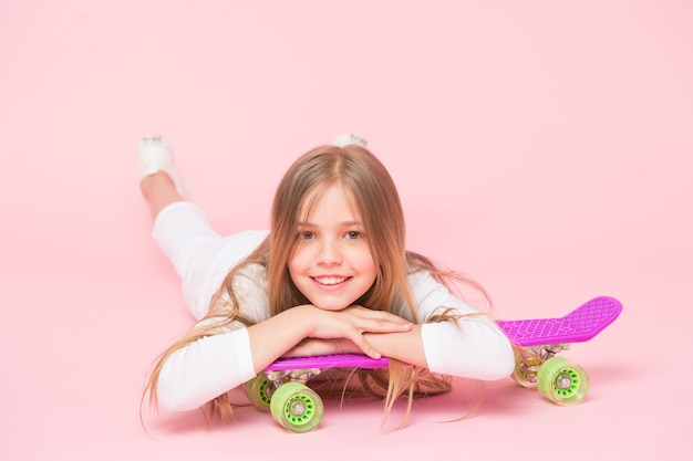 Menina na moda. Cabelo comprido de criança adorável criança adora passeio de penny board. Ande de penny board e faça truques. A menina gosta de andar de skate. Estilo de vida ativo. Menina se divertindo com fundo rosa de penny board.