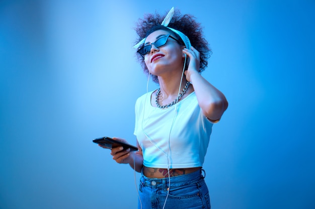 Menina na moda com cachos afro ouve e curte música com fones de ouvido em luz de néon azul, modelo estilo hip hop dança e relaxa