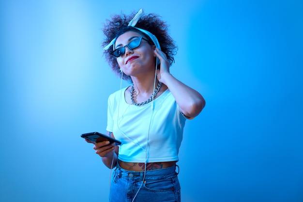 Menina na moda com cachos afro ouve e curte música com fones de ouvido em luz de néon azul, modelo estilo hip hop dança e relaxa