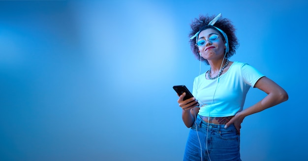 Menina na moda com cachos afro ouve e curte música com fones de ouvido em luz de néon azul