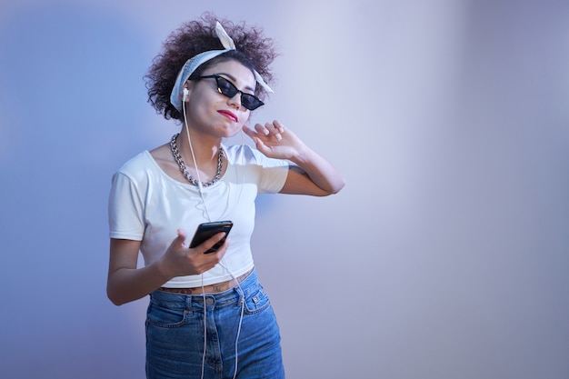 Menina na moda com cachos afro usando smartphone e fones de ouvido com espaço de cópia