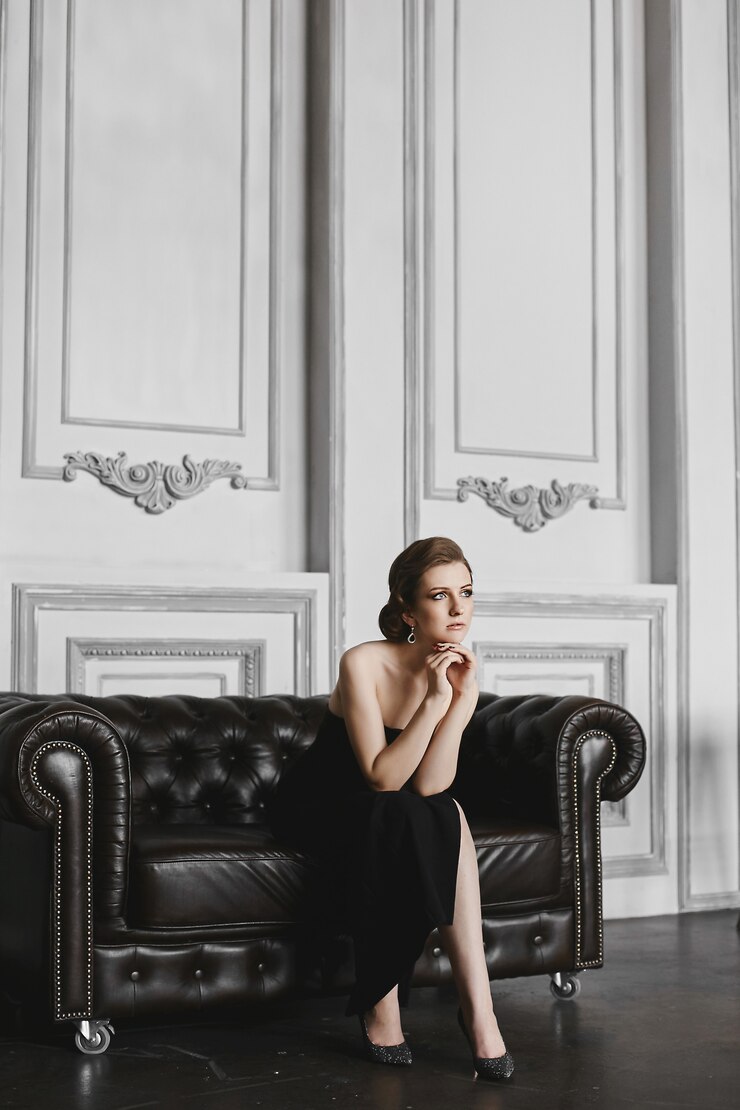 Foto menina na moda e elegante modelo morena com maquiagem suave e brincos grandes witj, vestido longo preto, senta-se no sofá vintage e posando no interior de luxo