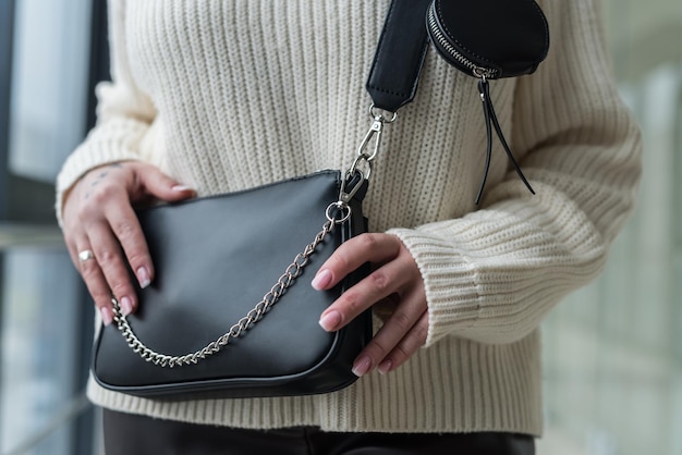 Menina na moda em um suéter aconchegante e quente mostra uma bolsa de couro preta elegante. O conceito de acessórios elegantes