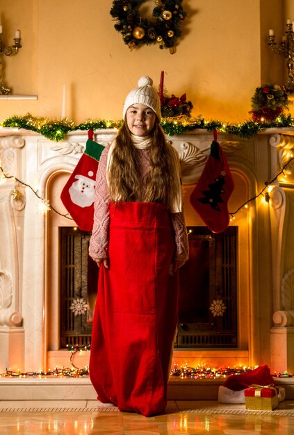 Menina na sacola vermelha do Papai Noel na sala de estar ao lado da lareira