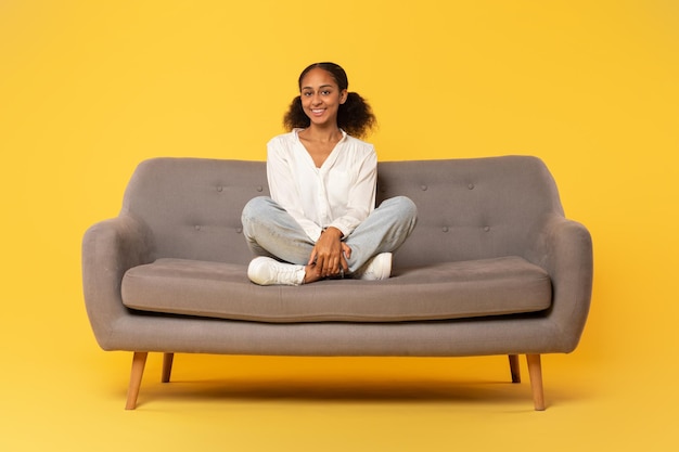 Foto menina negra feliz sentada cruzando as pernas no sofá fundo amarelo