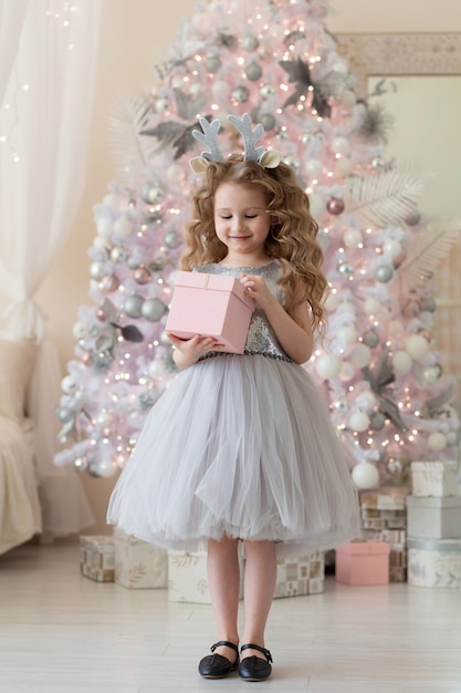Menina no aro com chifres de veado, com caixa de presente perto da árvore de Natal branca.