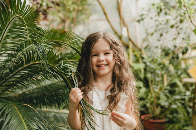 Menina no jardim botânico, uma garota de vestido branco ri perto de folhas de palmeira