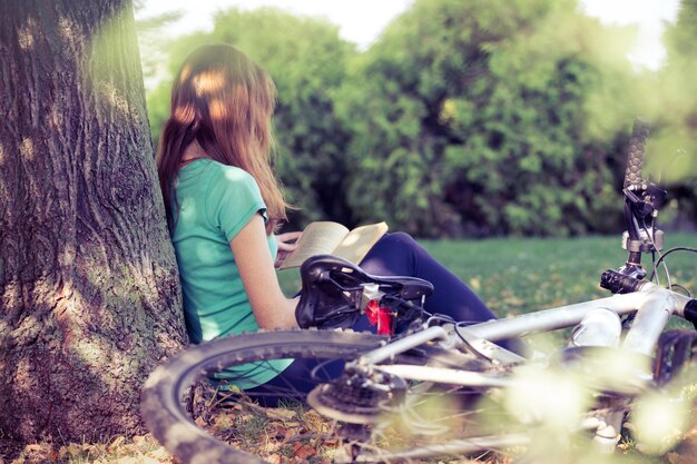 Menina no parque fica sob uma árvore e lê um livro. ao lado dela está uma bicicleta e uma mochila