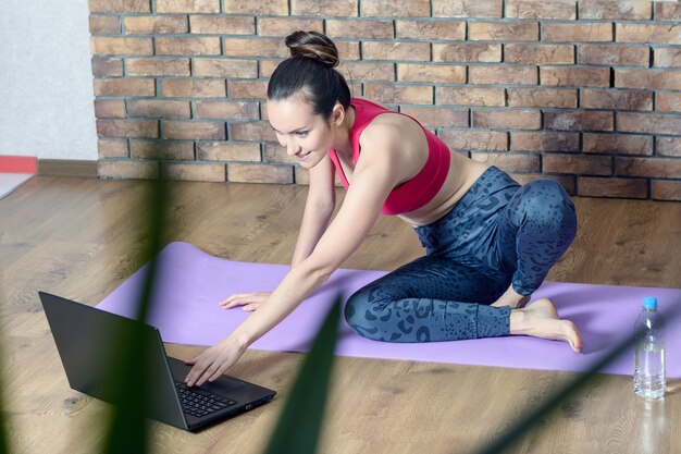 menina no sportswear faz pausa de exercícios enquanto está sentado no chão e ver a rede social no laptop com garrafa de água