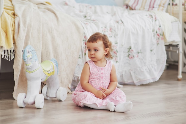Menina no vestido bonito localização na cama brincando com brinquedos em casa. Childroom vintage branco. Conceito de infância