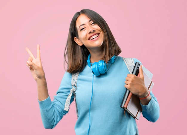 Menina nova do estudante com camiseta azul e fones de ouvido que sorri e que mostra o sinal da vitória.