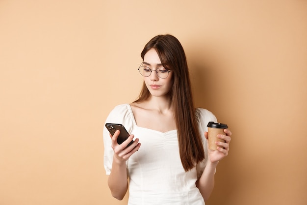 Menina ocupada de óculos lendo mensagem no celular e tomando café para viagem, em pé sobre fundo bege