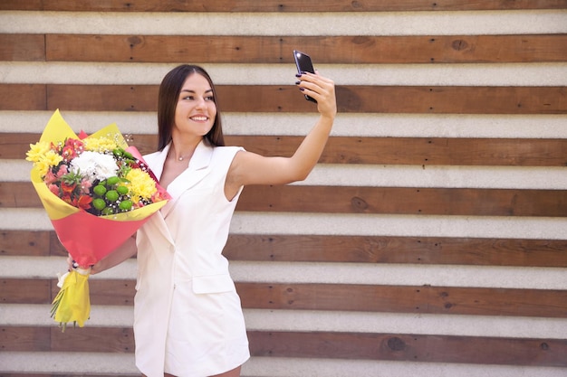 Menina oriental sorridente em roupas brancas com um buquê de flores tirando selfies no telefone