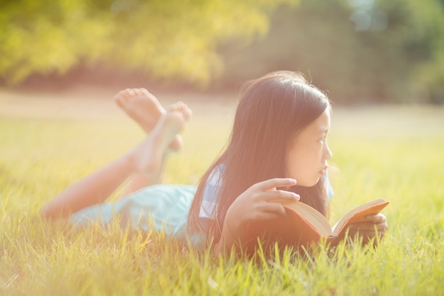 Menina pensativa, deitada na grama e livro de leitura no parque