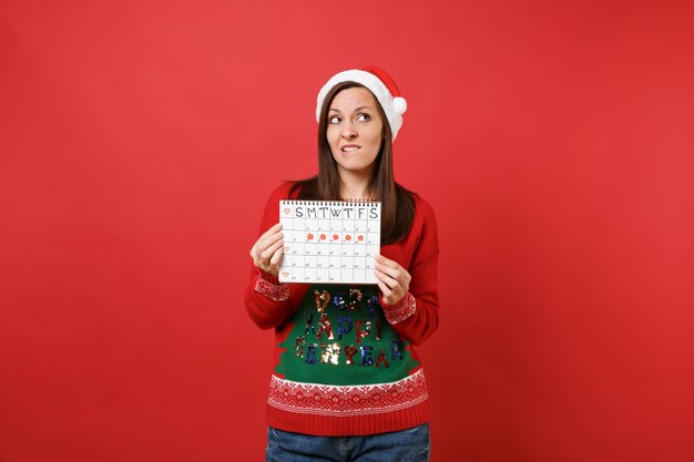 Menina pensativa do Papai Noel segurando o calendário de períodos femininos para verificar os dias de menstruação, isolados sobre fundo vermelho. Médica, saúde, ginecológica. Conceito de festa de feriado de celebração de ano novo feliz 2019.