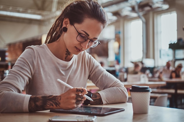 Foto menina pensativa elegante está esboçando no bloco de notas digital enquanto está sentado no café com uma xícara de café.