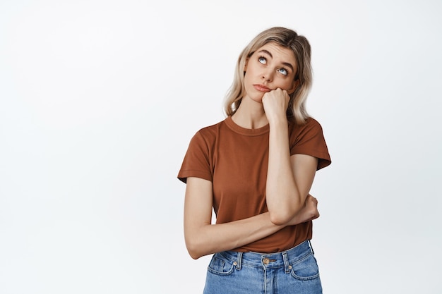 Menina pensativa, olhando para cima com a expressão do rosto entediado, em pé em uma camiseta marrom e jeans em branco.