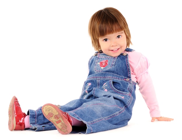 Foto menina pequena com macacão jeans, sentada no chão e sorrindo sobre uma parede branca. conceito de estilo de vida de lindas crianças felizes