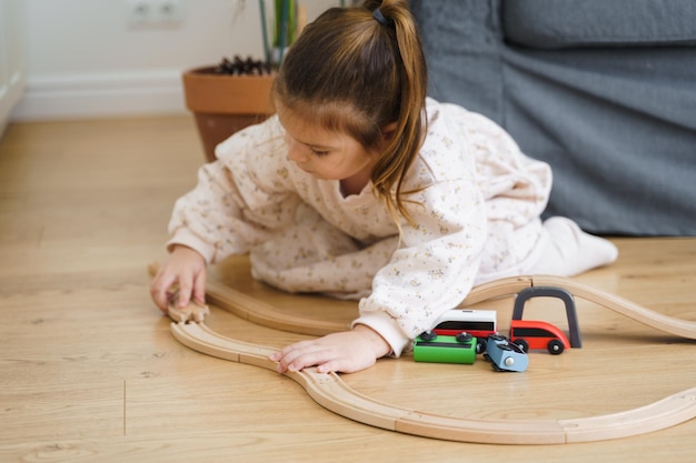 Menina pequena de vestido branco brinca com um trem de madeira em casa, na sala de estar