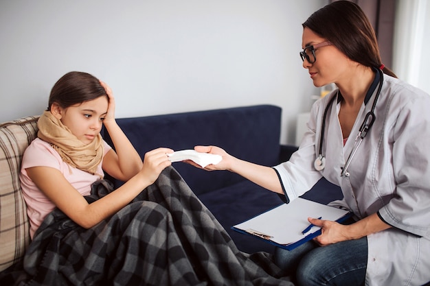 Menina pequena doente tirar tecido branco das mãos do médico. Ela olha para eles e segura uma mão na cabeça. Jovem simpática olhar paciente. Ela senta ao lado dela.