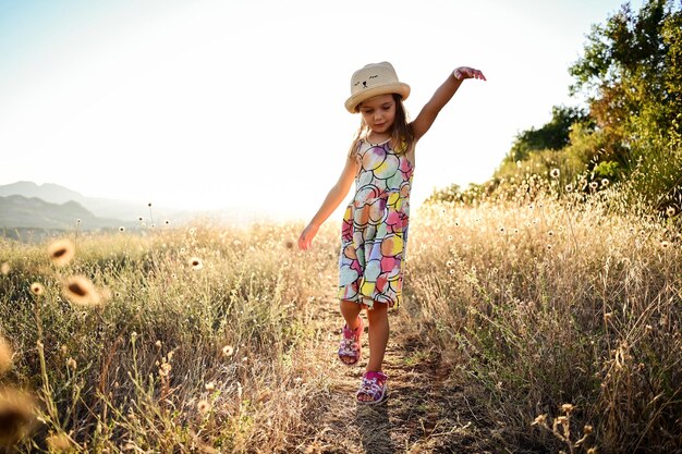 Foto menina pequena em um vestido de verão dançando no campo durante o pôr do sol