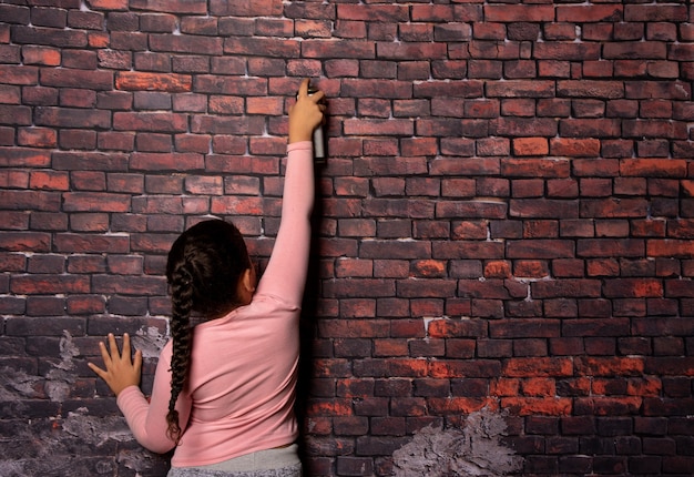 Foto menina pequena fazendo poses com um spray na frente de um cenário de parede de tijolo velho, fundo escuro, foco seletivo.