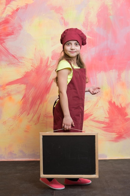 Menina pequena ou criança fofa com cara feliz no chapéu de chef vermelho e avental de cozinheiro segurando o quadro-negro em fundo abstrato colorido, cozinhando comida, copie o espaço